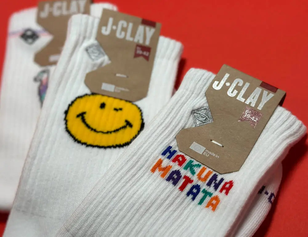 Drei Paar weiße J-CLAY Socken auf rotem Untergrund; eines hat ein gelbes Smiley-Gesicht, ein anderes hat HAKUNA MATATA in bunten Buchstaben, und das dritte zeigt ein kleines mehrfarbiges Muster.