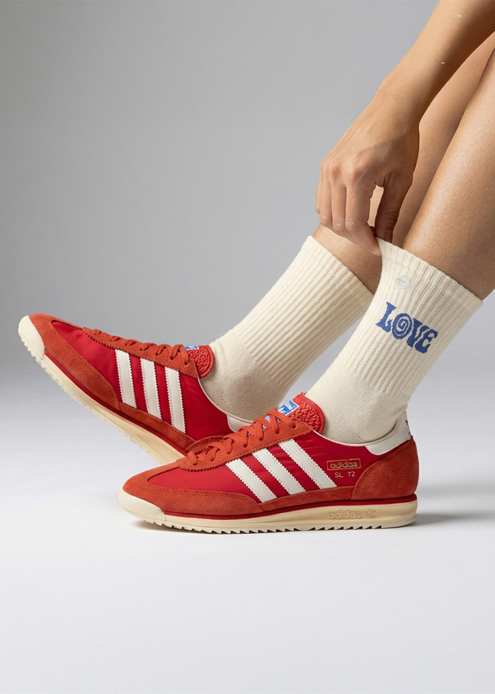 Socken kaufen - Eine Person in cremefarbenen Socken mit dem Wort LOVE in Blau und roten Adidas-Turnschuhen mit weißen Streifen, die mit gekreuzten Beinen auf einem grauen Hintergrund sitzt.