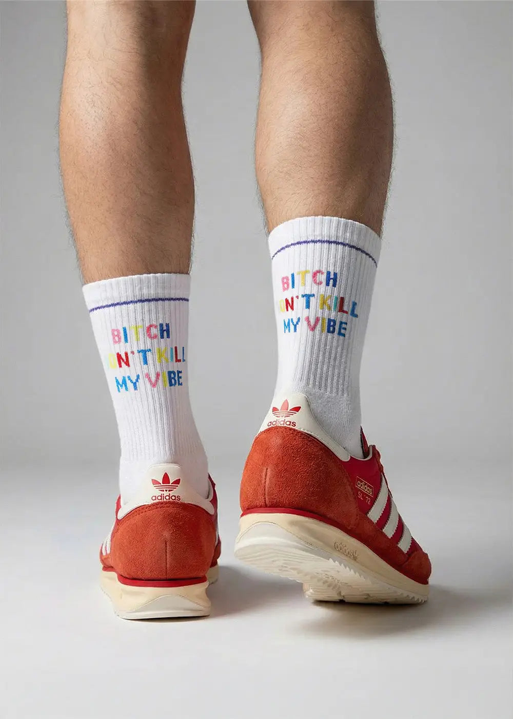 Eine Person, die weiße Socken mit der blauen Aufschrift Call me when youre lonely und blaue Adidas-Turnschuhe mit weißen Streifen trägt, beugt sich vor einem schlichten grauen Hintergrund.