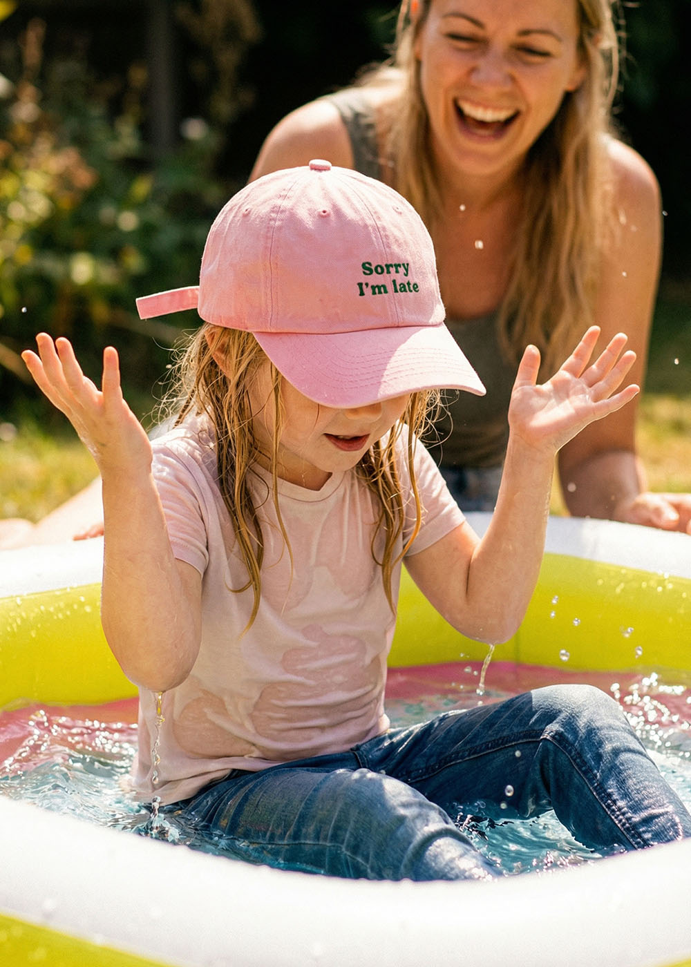 Ein kleines Kind mit rosa Mütze und Jeans planscht mit erhobenen Armen in einem kleinen aufblasbaren Pool. Ein Erwachsener lächelt im Hintergrund, draußen an einem sonnigen Tag. Auf der Mütze steht "Sorry I'm late".