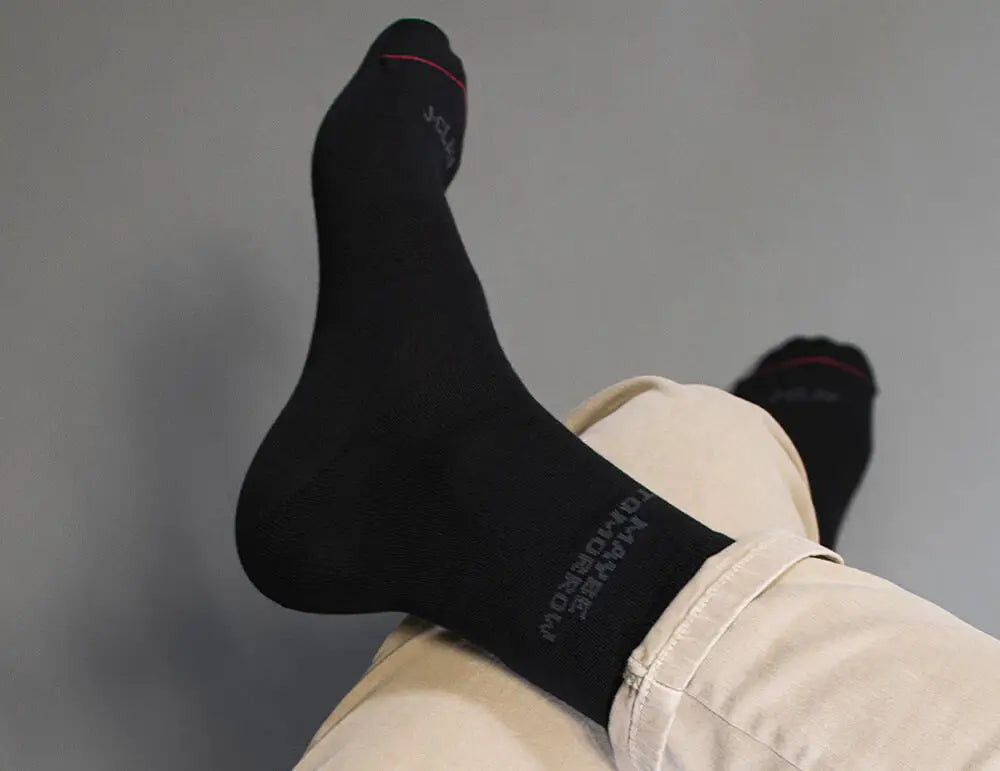 Eine Person in beiger Hose und schwarzen Socken mit übereinandergeschlagenen Beinen, die sich vor einem schlichten grauen Hintergrund ausruht. Die Socken haben einen schwachen Text und einen dünnen roten Streifen in der Nähe der Zehen.