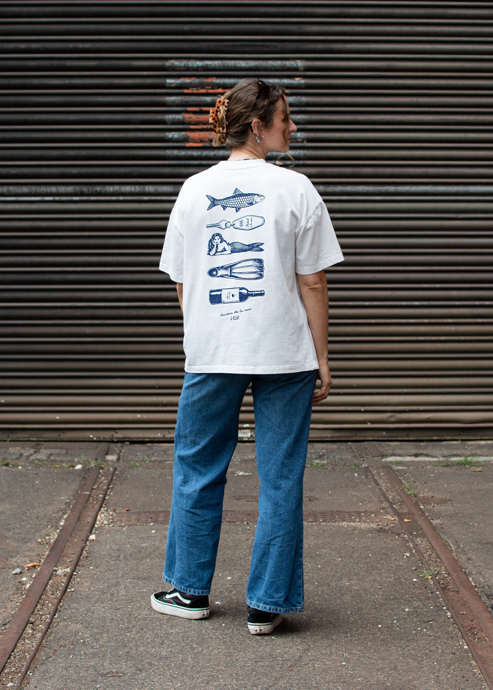 Eine Person in einem „J.Clay Ocean Kiss“-T-Shirt mit blauen Fisch- und Besteckmotiven steht auf einer Straße.