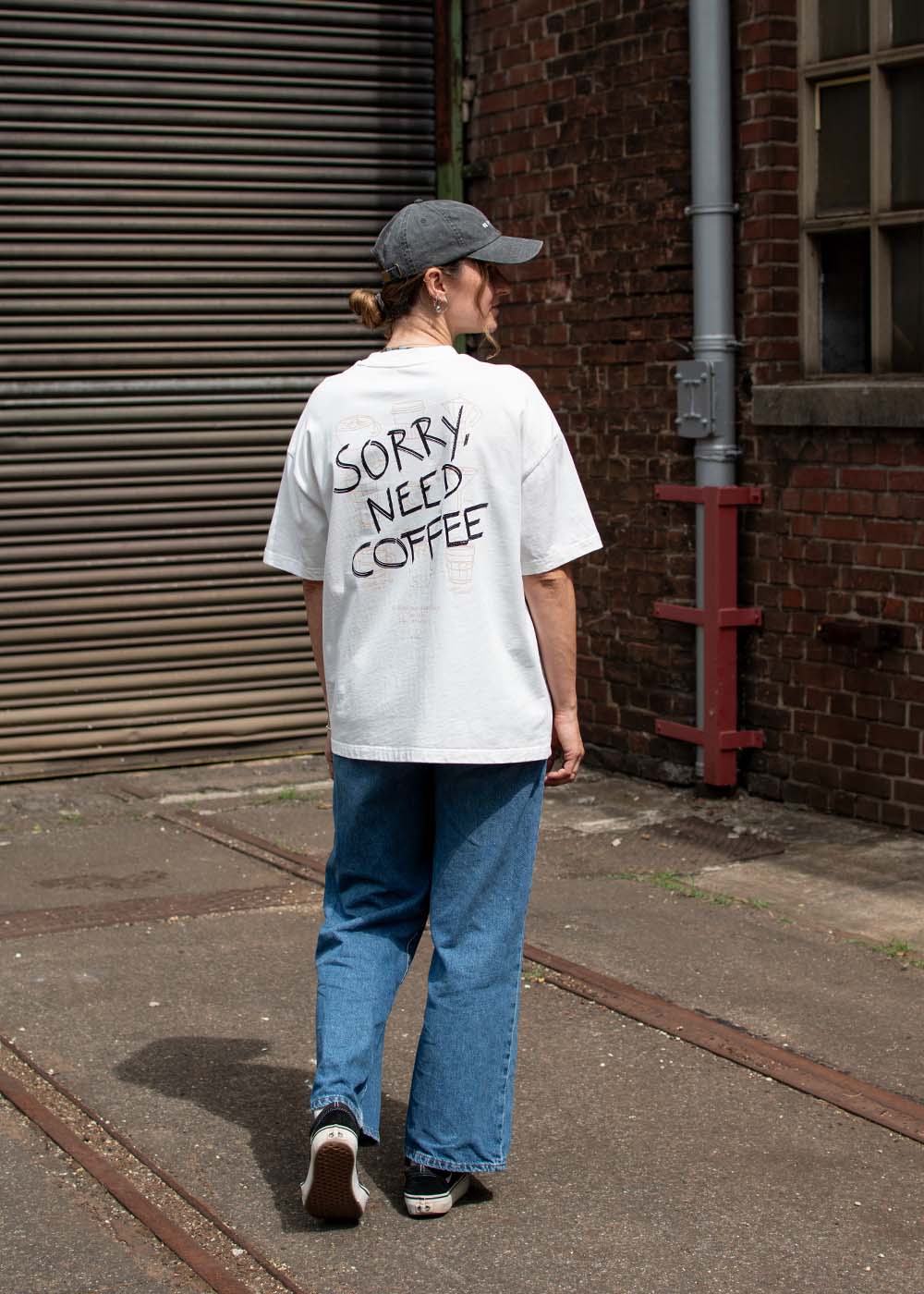 Eine Person in Jeans und Mütze trägt draußen ein T-Shirt von J.Clay mit der Aufschrift „Sorry Need Coffee“ in Unisex-Relaxed-Fit.