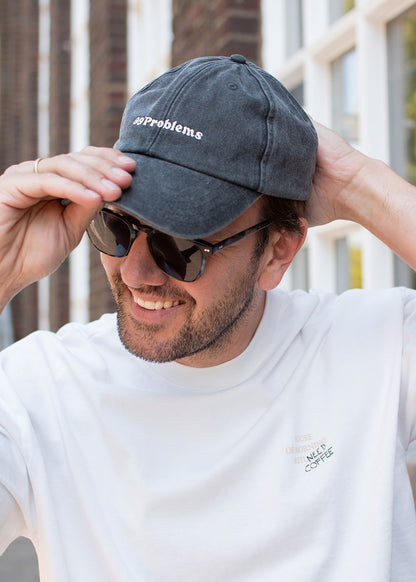 Ein Mann mit Sonnenbrille und weißem Hemd rückt draußen seine schwarze J.Clay-Kappe mit der Aufschrift „99 Problems“ zurecht.