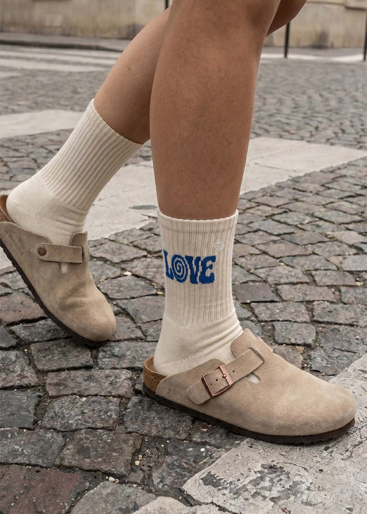Person mit J.Clay Boho Love-Socken und beigen Clogs auf einer Kopfsteinpflasterstraße.