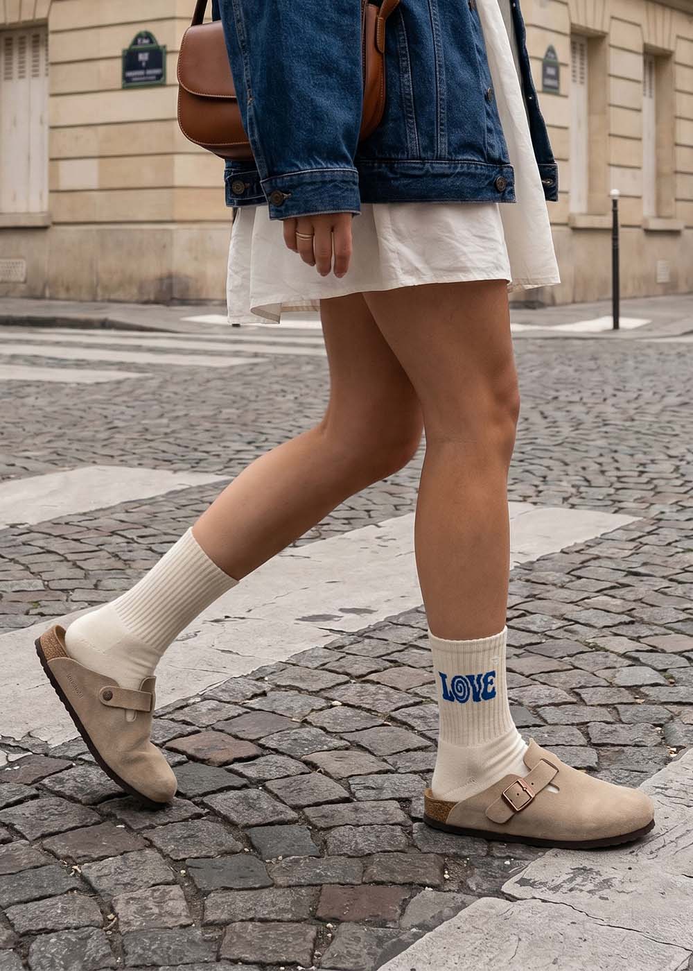 Person in weißem Kleid, Denim-Jacke und J.Clay boho love Socken spaziert.