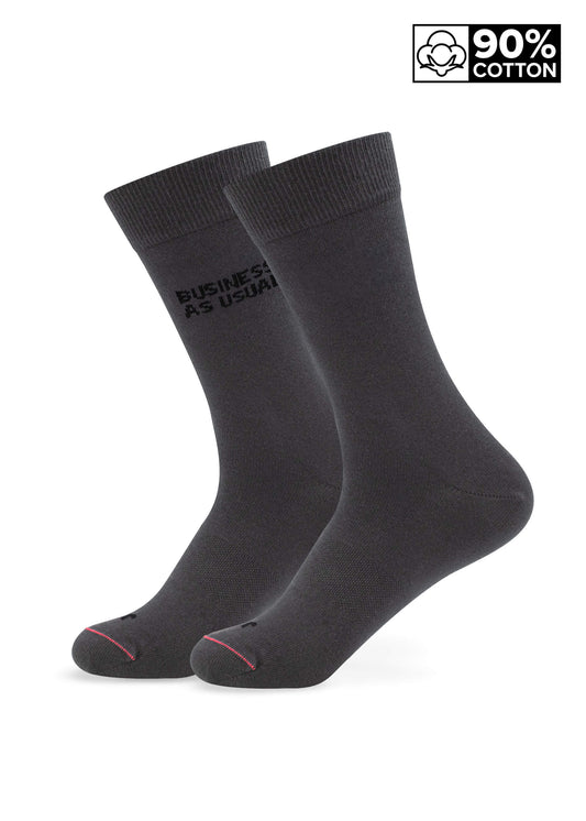 Die beiden schwarzen Crew-Business-Socken von J.Clay tragen den Text "BUSINESS AS USUAL" an der Seite und stehen aufrecht. Ein Ecketikett hebt 90% Baumwolle mit einem Baumwollsymbol hervor und kombiniert Komfort und Stil.