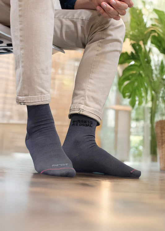 Eine Person in beiger Hose und schwarzen J.Clay-Business-Socken - eine davon mit der Aufschrift "BUSINESS AS USUAL" - sitzt auf einem Stuhl mit einer Grünpflanze und einem verschwommenen Fenster im Hintergrund.