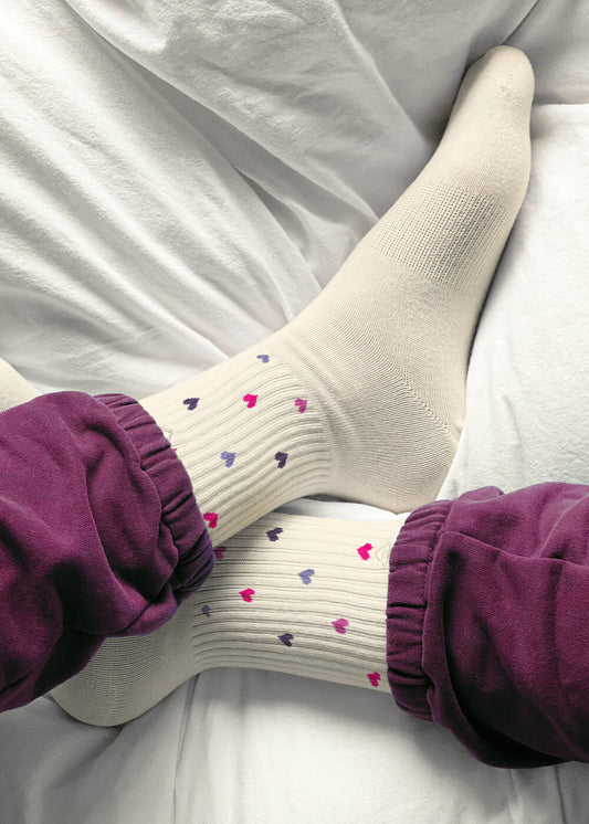 Eine Person, die cremefarbene Tennissocken von J.Clay mit kleinen bunten Herzmustern und eine lila Hose trägt, liegt auf einem weißen Bett, die Beine an den Knöcheln gekreuzt.