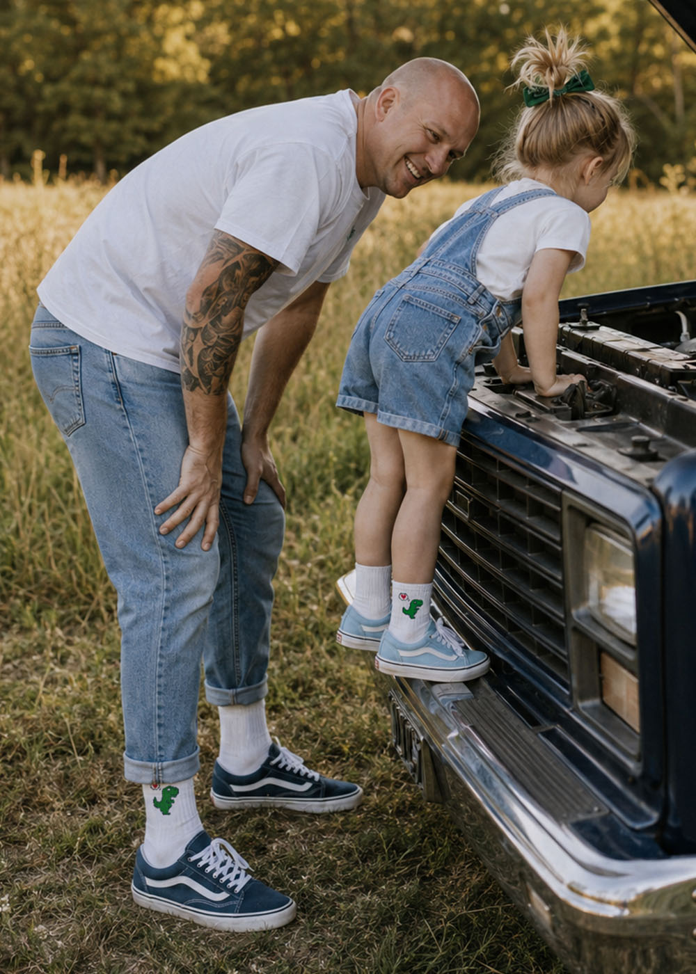 Ein kahlköpfiger Mann und ein Mädchen aus „Dino Love – Fam Pack“ von J.Clay schauen auf einem Feld unter die Motorhaube eines Lastwagens.