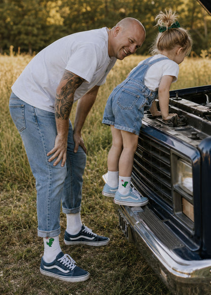 Ein kahlköpfiger Mann und ein Mädchen aus „Dino Love – Fam Pack“ von J.Clay schauen auf einem Feld unter die Motorhaube eines Lastwagens.