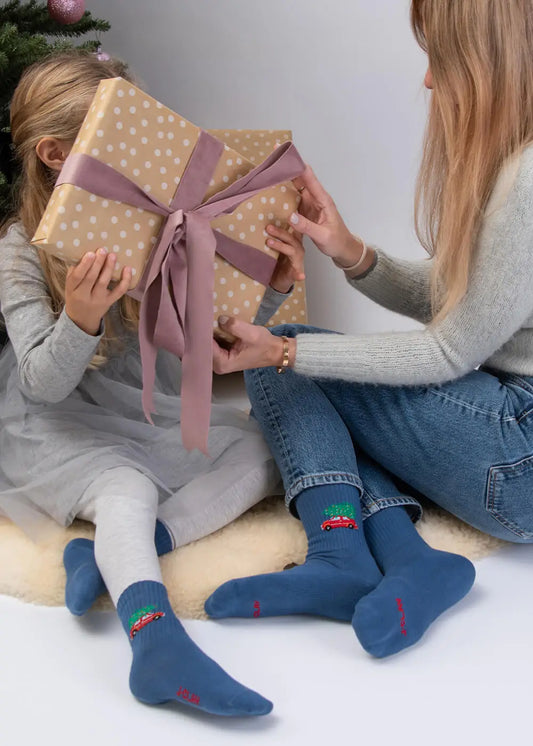 Kind mit dem Song „Driving Home for Christmas – Kids“ von J.Clay, neben einem Erwachsenen in der Nähe des Weihnachtsbaums.