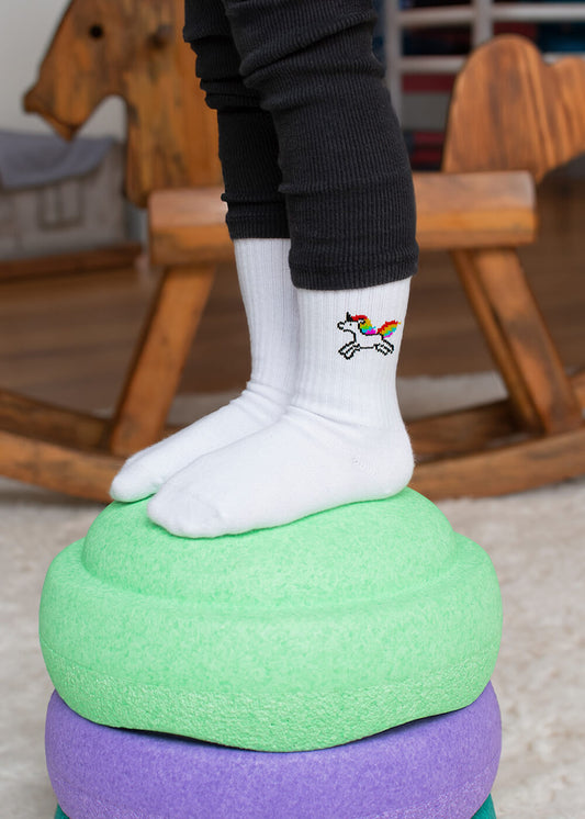 Ein Kind in schwarzen Hosen steht auf bunten Schaumstoff-Trittsteinen und trägt weiße J.Clay-Socken mit einem kleinen, aufgestickten Regenbogen-Einhorn. Im Hintergrund ist ein hölzernes Schaukelpferd zu sehen.