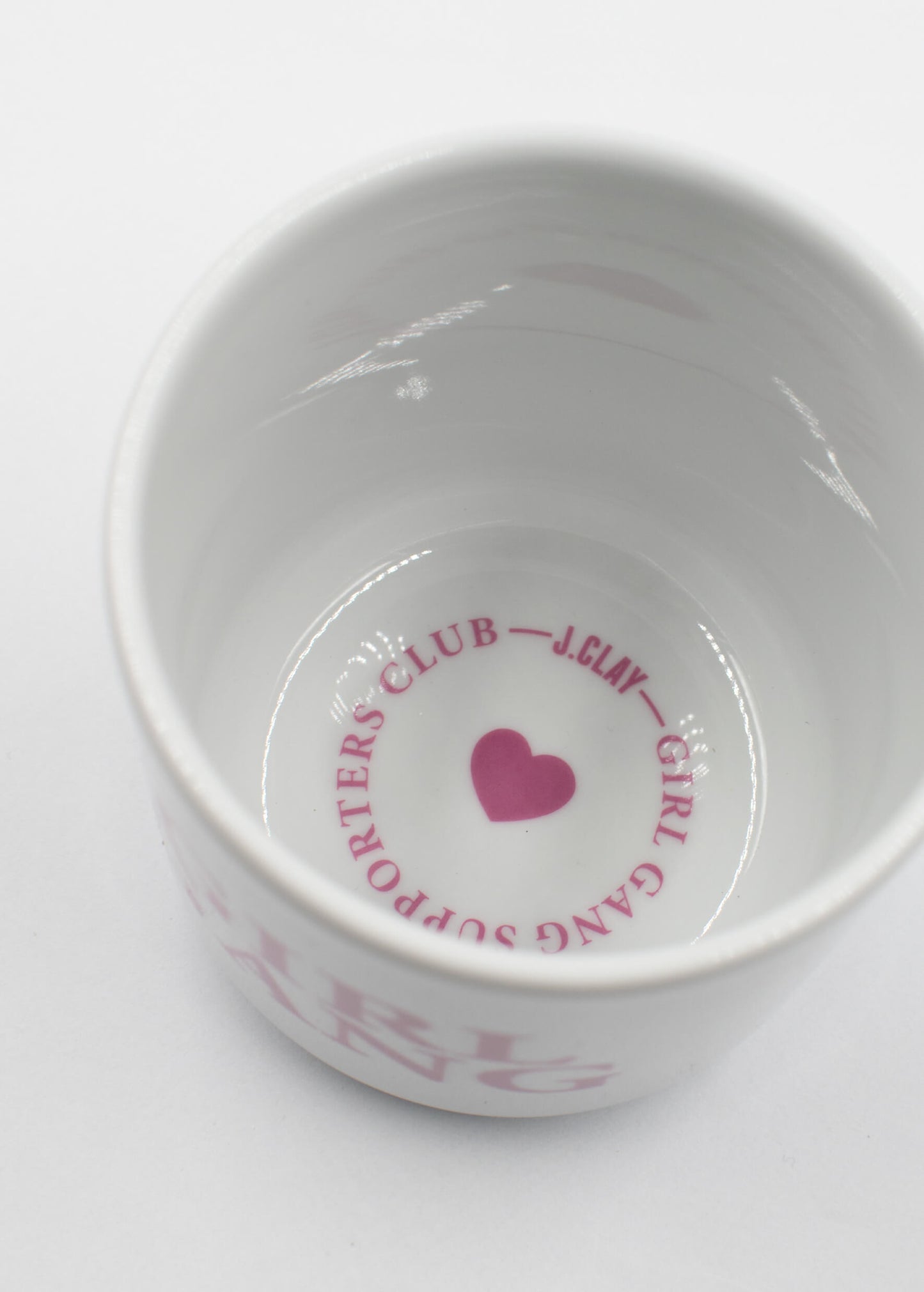 Eine weiße Keramik J.Clay Tasse ohne Henkel von oben gesehen, mit einem rosa Herz und "SUPPORTERS CLUB - J.Clay - GIRL GANG" in einem Kreis auf dem inneren Boden.