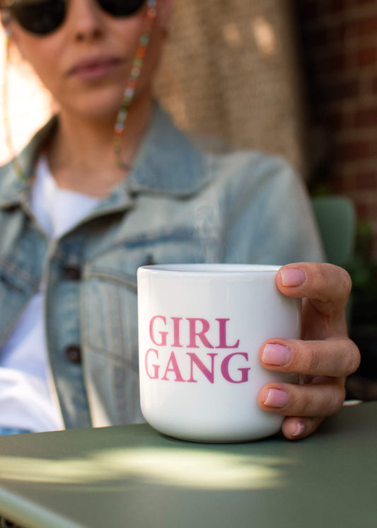 Eine Person in Jeansjacke und Sonnenbrille hält eine J.Clay Tasse ohne Henkel mit dem rosafarbenen Schriftzug "GIRL GANG" in der Hand. Der Hintergrund ist leicht unscharf, damit der Fokus auf der Hand und der Tasse bleibt.