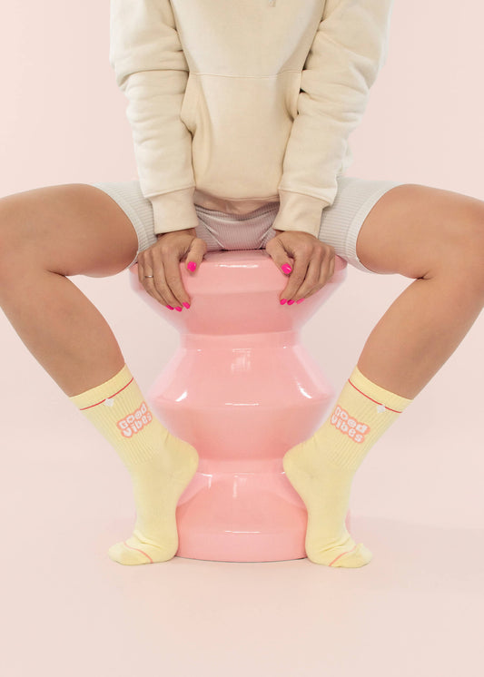 In einem beigen J.Clay-Kapuzenpulli, hellen Shorts und gelben J.Clay-Tennissocken mit "Good Vibes"-Schriftzug sitzt eine Person auf einem rosa Hocker, auf dem rosa Nägel zu sehen sind. Der pastellrosa Hintergrund unterstreicht die Stimmungs-Booster-Stimmung.