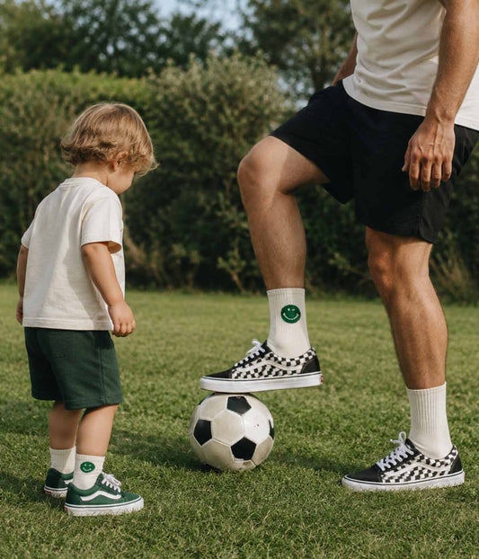 Kleinkind und Erwachsener in Socken der Marke „J.Clay Green Smile – Fam Pack“, die draußen in Turnschuhen Fußball spielen.