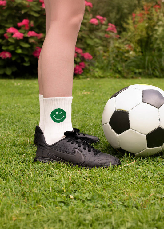 Eine Person in schwarzen J.Clay Turnschuhen und weißen Kindersocken mit grünem Smile-Motiv steht auf dem Rasen zwischen einem schwarz-weißen Fußball, rosa Blumen und Grünzeug - ideal für ein fröhliches 3er Pack von J.Clay.