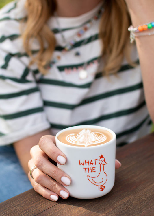 Frau mit Latte in „J.Clay What the Duck“ – Tasse mit der Aufschrift „WHAT THE“ und einer Cartoon-Gans.