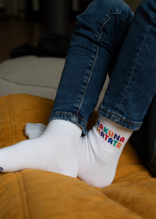 Eine Person in Bluejeans und J.Clay Kindersocken ab 5 Jahren mit buntem "Hakuna Matata"-Text sitzt im Schneidersitz auf einem gelben Kissen.