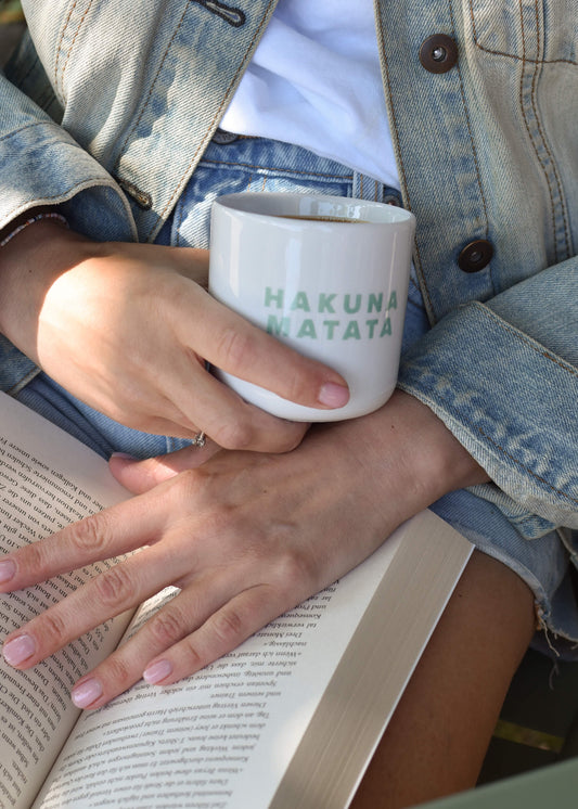 Jemand trägt Jeans-Shorts und eine Jacke, hält eine J.Clay HAKUNA MATATA Tasse ohne Henkel in der Hand und stützt sich auf ein aufgeschlagenes Buch - ein friedlicher Moment, der mit dem Hakuna Matata Pack oder Geschenkset von J.Clay noch schöner wird.