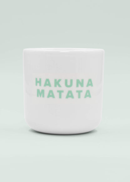 Eine weiße Keramik-Tasse ohne Henkel von J.Clay mit der Aufschrift "HAKUNA MATATA" in hellgrünen Großbuchstaben auf der Vorderseite vor einem hellen Hintergrund.