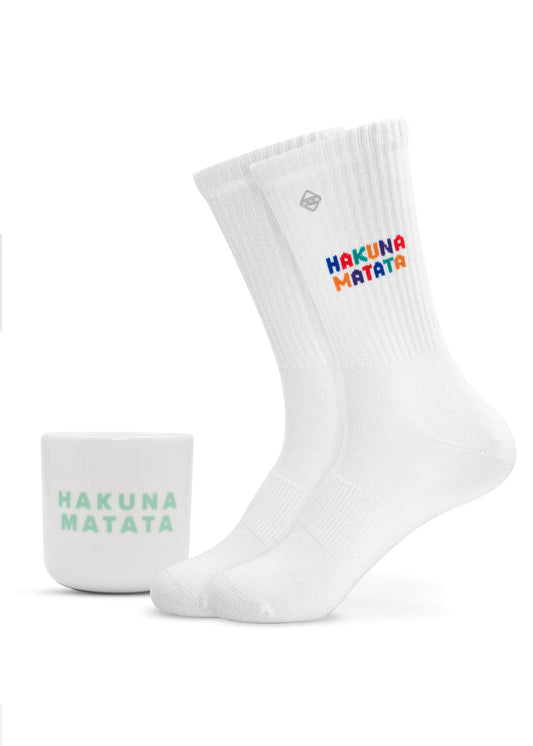Fröhliches J.Clay-Geschenkset: weiße Socken mit buntem "Hakuna Matata"-Schriftzug und -Logo, gepaart mit einer grifflosen Tasse mit grünem "Hakuna Matata"-Aufdruck - perfekt als Hakuna Matata-Pack vor einem schlichten weißen Hintergrund.