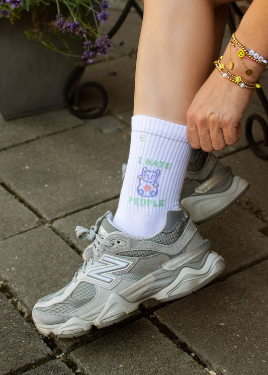 Eine Person in grauen Turnschuhen und weißen Tennissocken von J.Clay mit einem Cartoon-Bären und dem Text "I HATE PEOPLE" sitzt auf einer Steinterrasse, bunte Perlenarmbänder am Handgelenk und lila Blumen im Hintergrund.