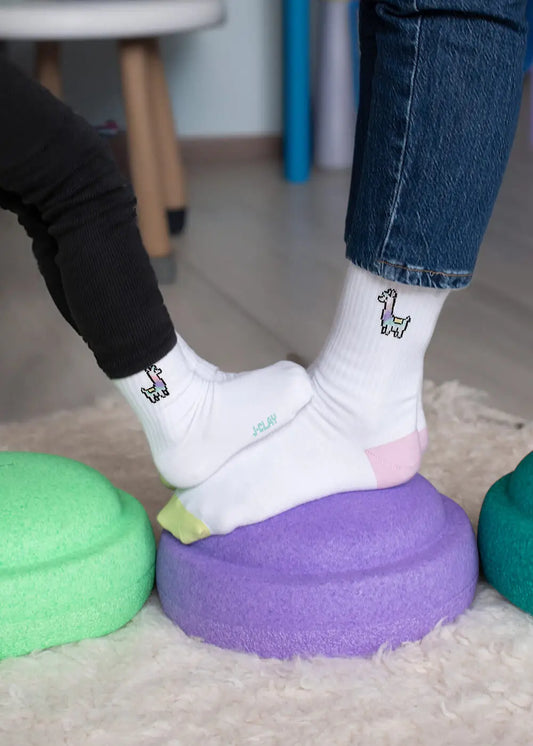 Zwei Personen, die J.Clay-Socken im Partnerlook mit Cartoon-Lama-Motiven tragen, stehen auf grünen und lila strukturierten Kissen auf einem hellen Holzboden. Nur ihre Beine und Füße sind zu sehen, was den lustigen, passenden J.Clay-Sockenstil unterstreicht.