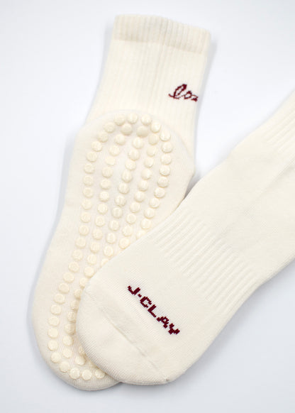 Zwei weiße J.Clay Love Grip Socken mit Eco Grip Sohlen und rotem J.Clay Text auf einem.