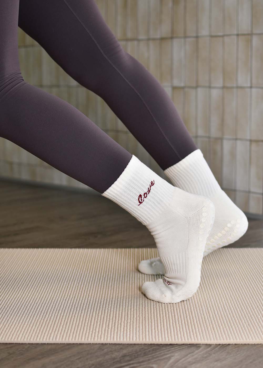 Eine Person in weißen J.Clay Love Grip-Socken und dunklen Leggings steht auf einer beigen Matte für Reformer Pilates.