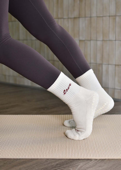 Eine Person in weißen J.Clay Love Grip-Socken und dunklen Leggings steht auf einer beigen Matte für Reformer Pilates.