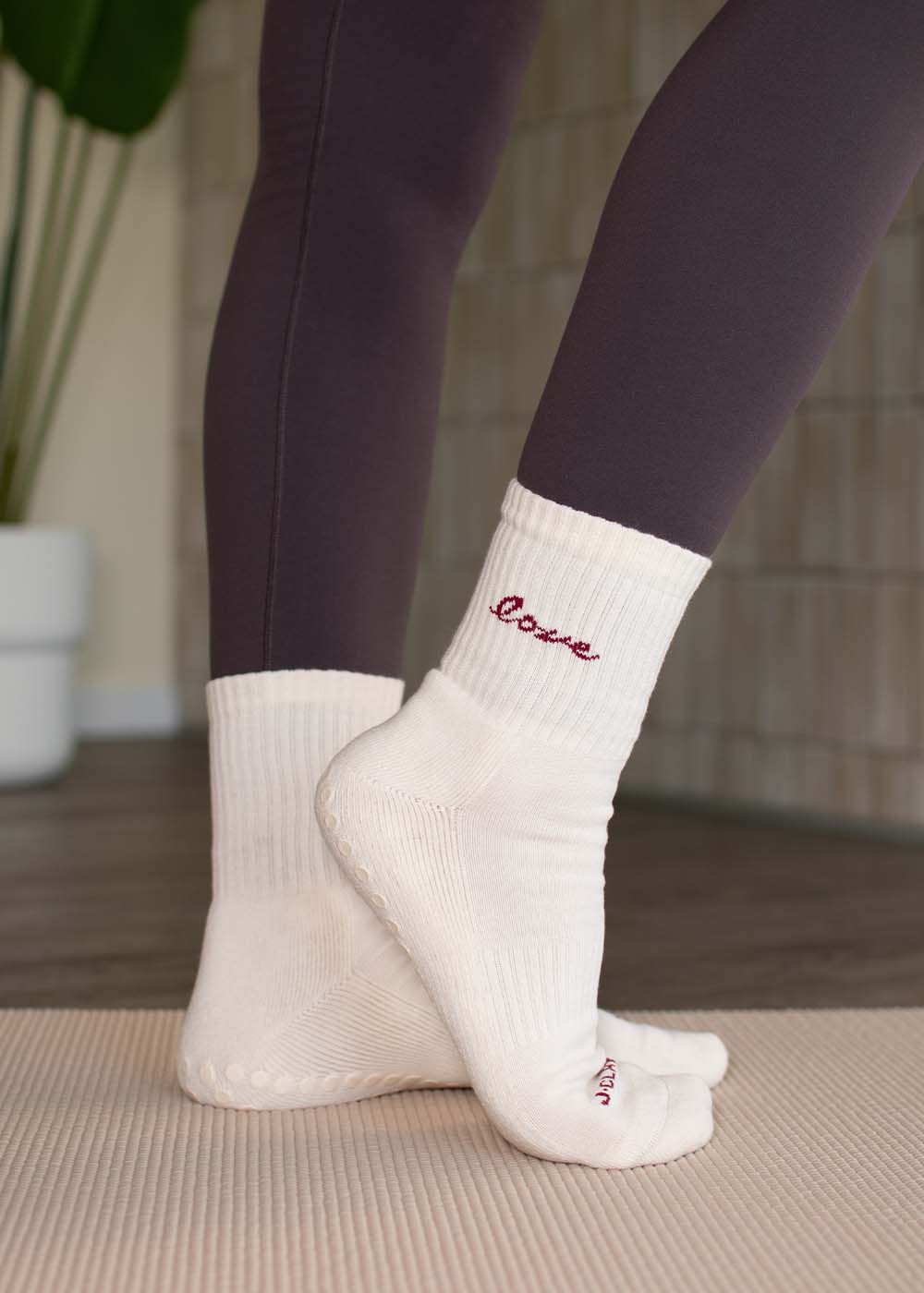 Person, die J.Clay Love Grip Socken und dunkle Leggings trägt und auf einer Yogamatte in einem Raum steht.