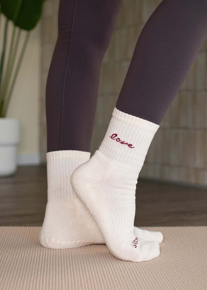Person, die J.Clay Love Grip Socken und dunkle Leggings trägt und auf einer Yogamatte in einem Raum steht.