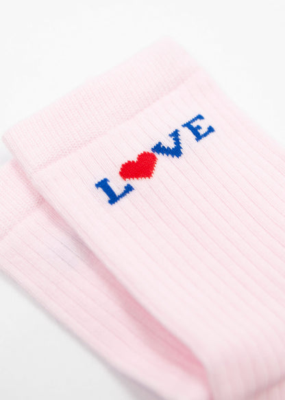 Eine Nahaufnahme von rosa gerippten J.Clay Damen Socken mit "LOVE", das in der Nähe des oberen Randes gestickt ist - das O ist ein rotes Herz, die anderen Buchstaben sind blau. Die Socken liegen ordentlich gefaltet auf einer weißen Fläche.