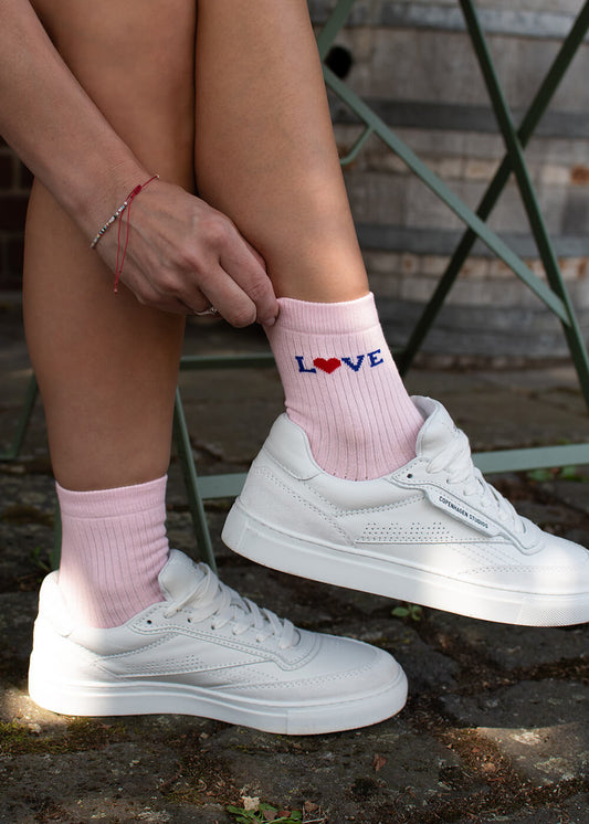 Eine Person in weißen Turnschuhen und J.Clay Rosa Socken - mit "LOVE" und einem roten Herz - zieht eine Socke hoch, während sie draußen auf einem grünen Metallstuhl über Steinpflaster sitzt. Diese Damen Socken bringen Blossom Love Charme zu jedem Freizeitoutfit.