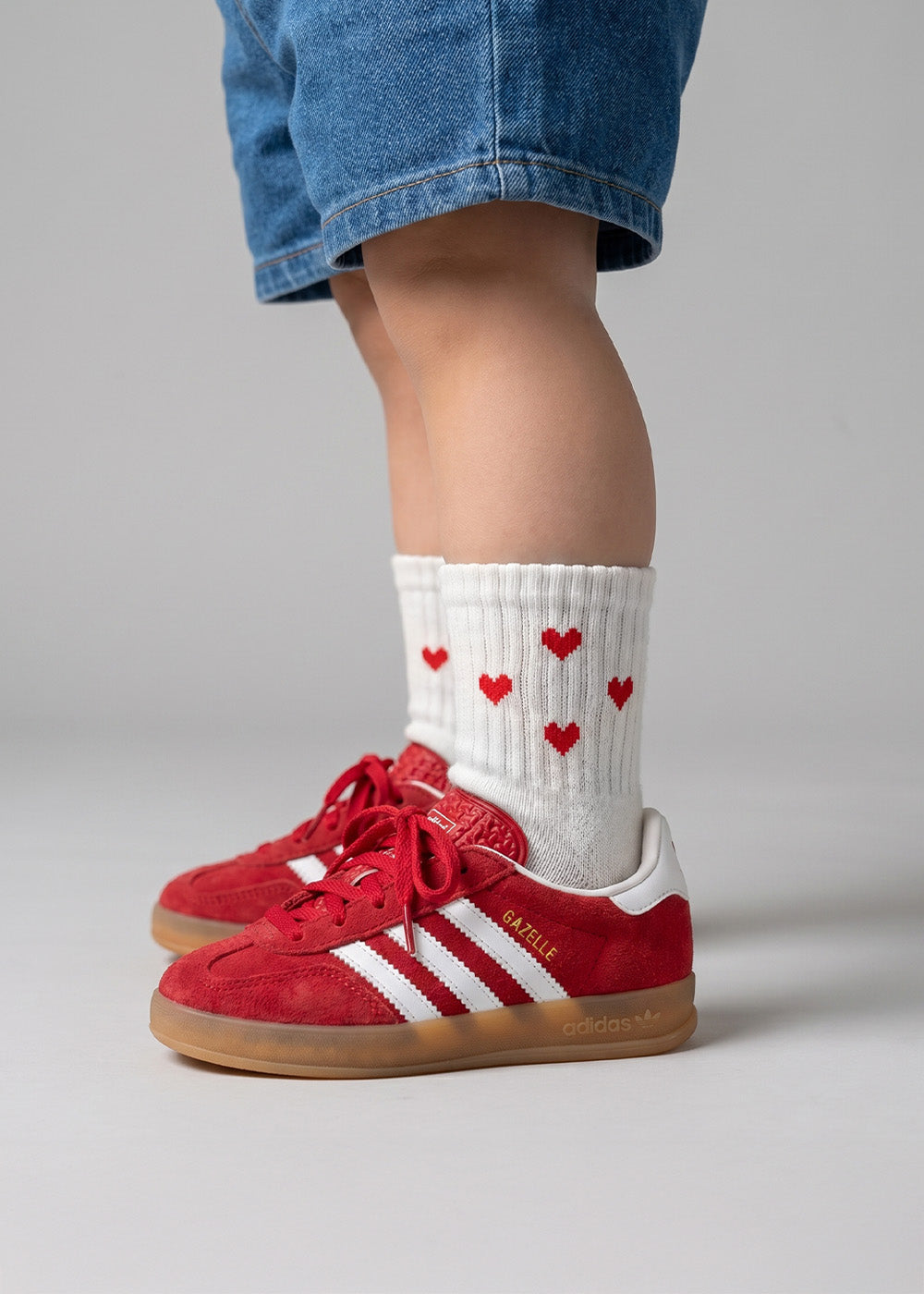 Ein Kind in blauen Shorts, J.Clay Red Hearts – Minisocken und roten Turnschuhen, stehend.