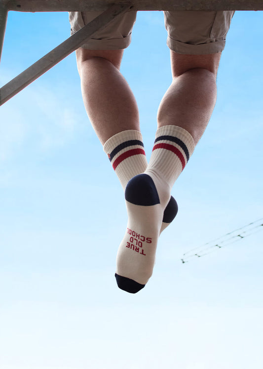 Eine Person in beigen Shorts und J.Clay Tennissocken - cremefarbene Socken mit roten und marineblauen Streifen - sitzt mit baumelnden Beinen da und zeigt ihre Fußsohlen, die vor dem blauen Himmel TRUE OLD SCHOOL sagen.