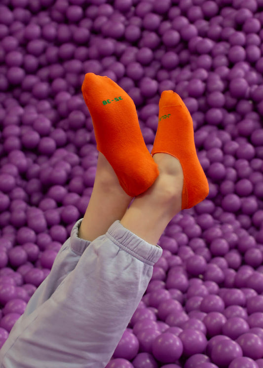 Eine Person, die leuchtend orangefarbene J.Clay-Socken und eine helllavendelfarbene Hose trägt, hebt ihre Beine über eine mit lila Plastikbällen gefüllte Ballgrube, selbstbewusst, als hätte sie rutschfeste Füßlinge an.