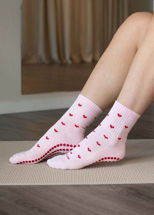 Person, die J.Clay Red Hearts Grip Socken trägt und auf einer Matte auf einem Holzboden sitzt.