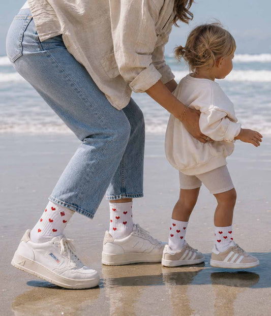 Ein Erwachsener und ein Kind in „J.Clay Red Hearts – Fam Pack“ stehen gemeinsam an einem Sandstrand in der Nähe der Wellen.