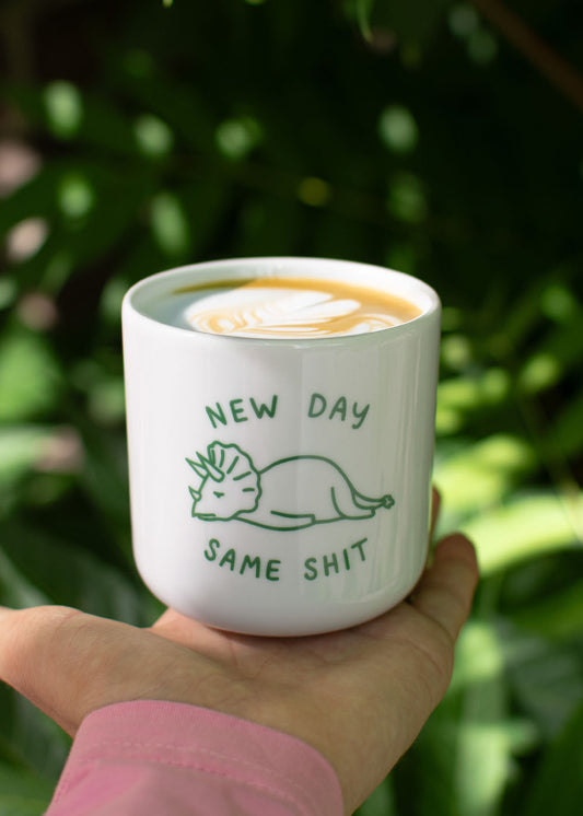 J.Clay New Day – Tasse: weiße Kaffeetasse mit Dinosaurier, Latte-Art und dem Schriftzug „NEW DAY SAME SHIT“.