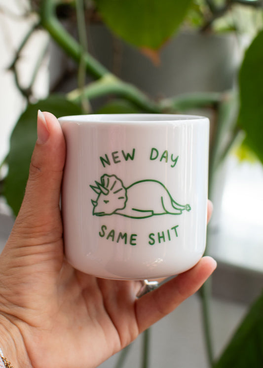Eine Hand, die eine Tasse von J.Clay „New Day“ mit einem Cartoon-Dino-Motiv und einem sarkastischen grünen Schriftzug hält.