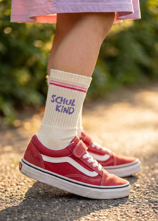 Kind in J.Clay Schulkind - Orange Purple Kids Socken steht in roten Turnschuhen auf einem sonnenbeschienenen Weg.