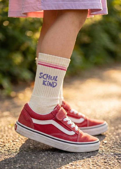 Kind in J.Clay Schulkind - Orange Purple Kids Socken steht in roten Turnschuhen auf einem sonnenbeschienenen Weg.