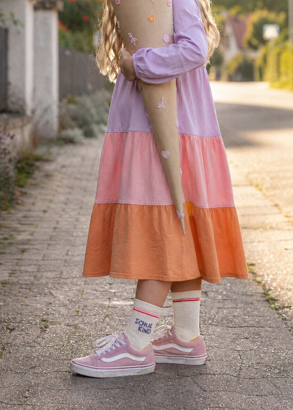 Ein Kind in einem J.Clay Schulkind - Orange Purple Kids Kleid hält eine Tüte für den ersten Schultag.