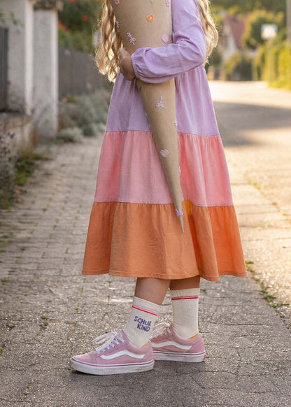 Ein Kind in einem J.Clay Schulkind - Orange Purple Kids Kleid hält eine Tüte für den ersten Schultag.