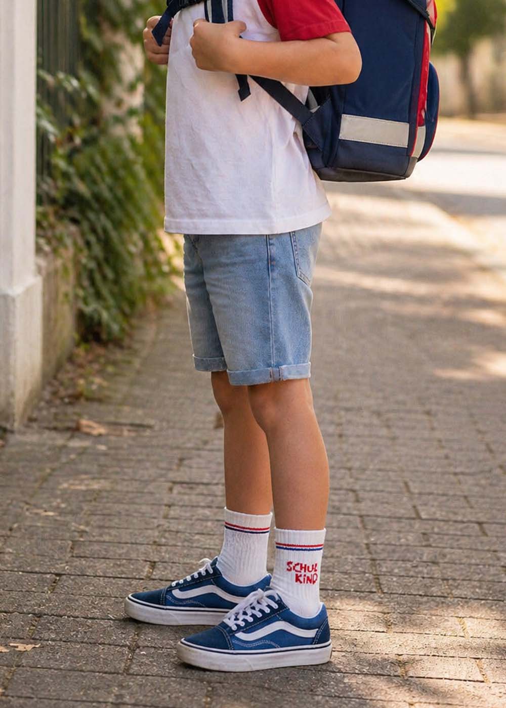 Kind in Shorts, Turnschuhen und J.Clay Schulkind - Red Blue Kids Socken steht mit einem Rucksack.