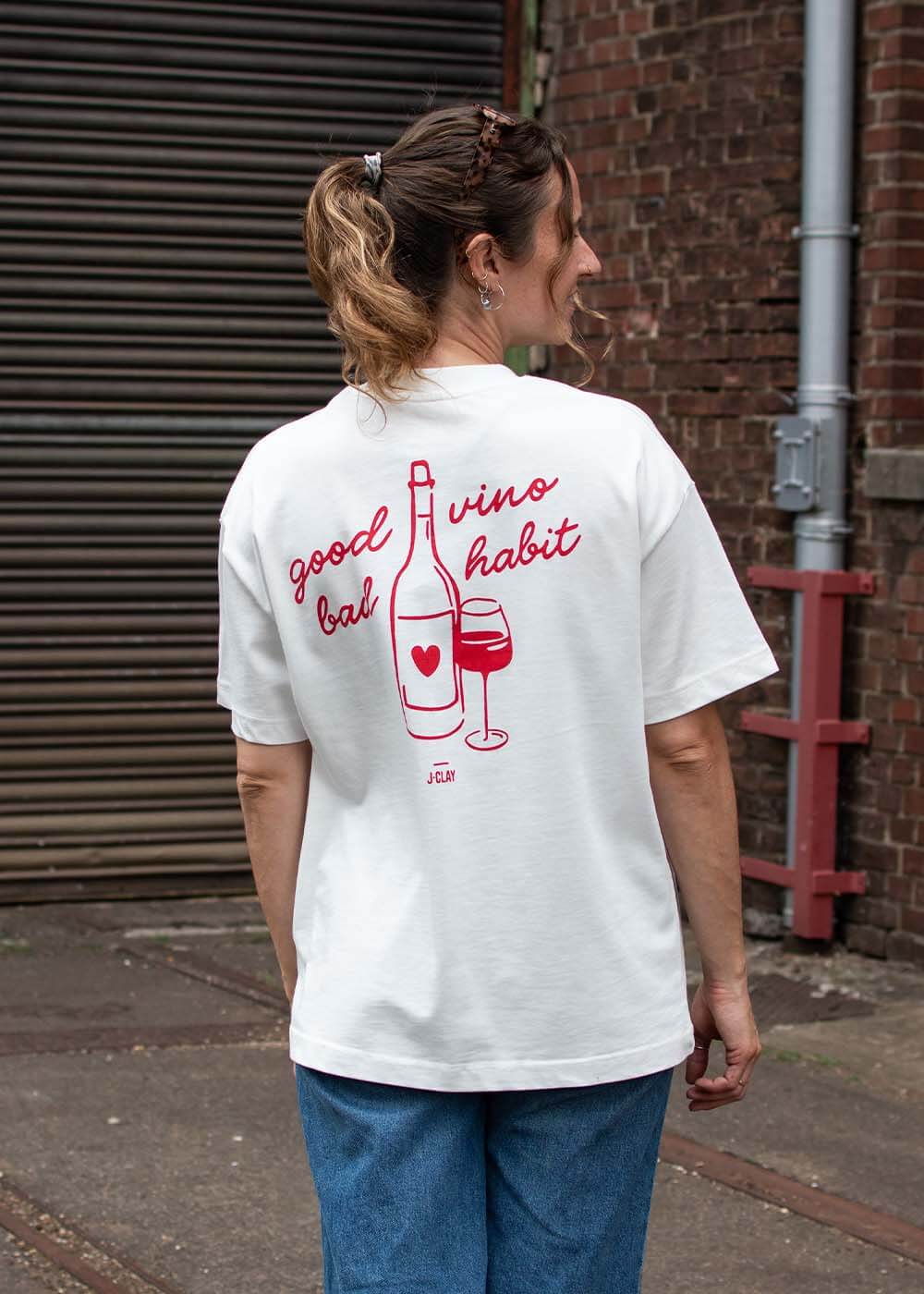 Frau in einem T-Shirt von J.Clay Good Vino mit Weinmotiv, draußen vor einer Backsteinmauer.