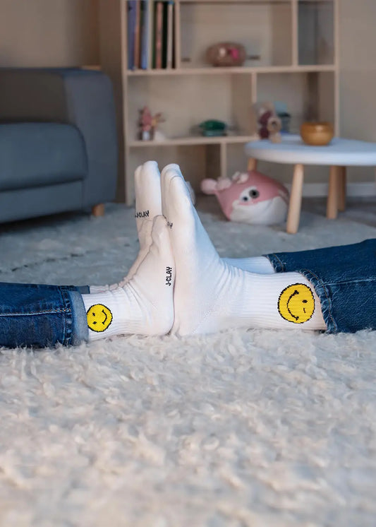 Zwei Personen, die J.Clay Mini Me Socken mit gelben Smiley-Gesichtern tragen, berühren sich mit den Füßen auf einem flauschigen weißen Teppich und schaffen so eine gemütliche Atmosphäre im Partnerlook in einem Wohnzimmer mit Bücherregalen und Kuscheltieren.